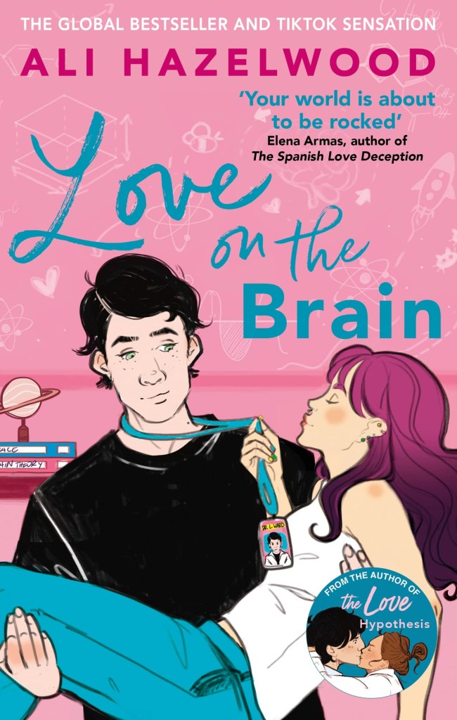 This is a cover image of Love on the Brain by Ali Hazelwood. The cover is pink, and shows a woman being held bridal style by a man. Behind the couple is a desk with books and planet diorama on it. The man wears a black t-shirt, he has black hair, and very green eyes. The woman in his arms tugs him towards her puckered lips by his lanyard. She has pink and purple hair, and is wearing jeans and a vest top. Her eyes are closed, and she is offering to kiss him.