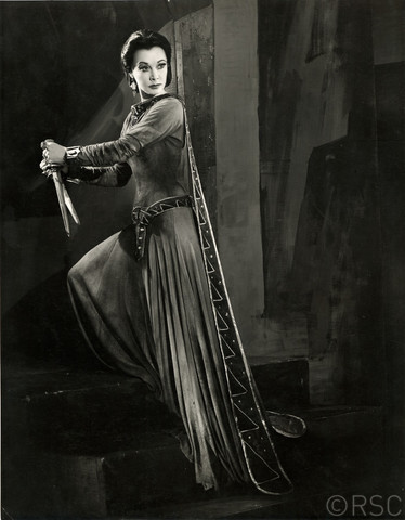 Macbeth, 1955: Lady Macbeth grasps the bloodied daggers Photo by Angus McBean. A beautiful dark haired woman, with a widow's peak, stands upon the stair. Her black hair is pulled into a bun at the base of her neck, casting her delicate, sharp features in sharp relief. She wears a long, largely plain dress, embellished at the neck, cuffs, waist by  intricate embroidery. A cape flows from the shoulders of the gown to sweep the floor. She looks very determined. In each hand she holds a dagger, they hover at chest height, clenched in sure hands. She is unpractised as an assassin, but certain of her action.